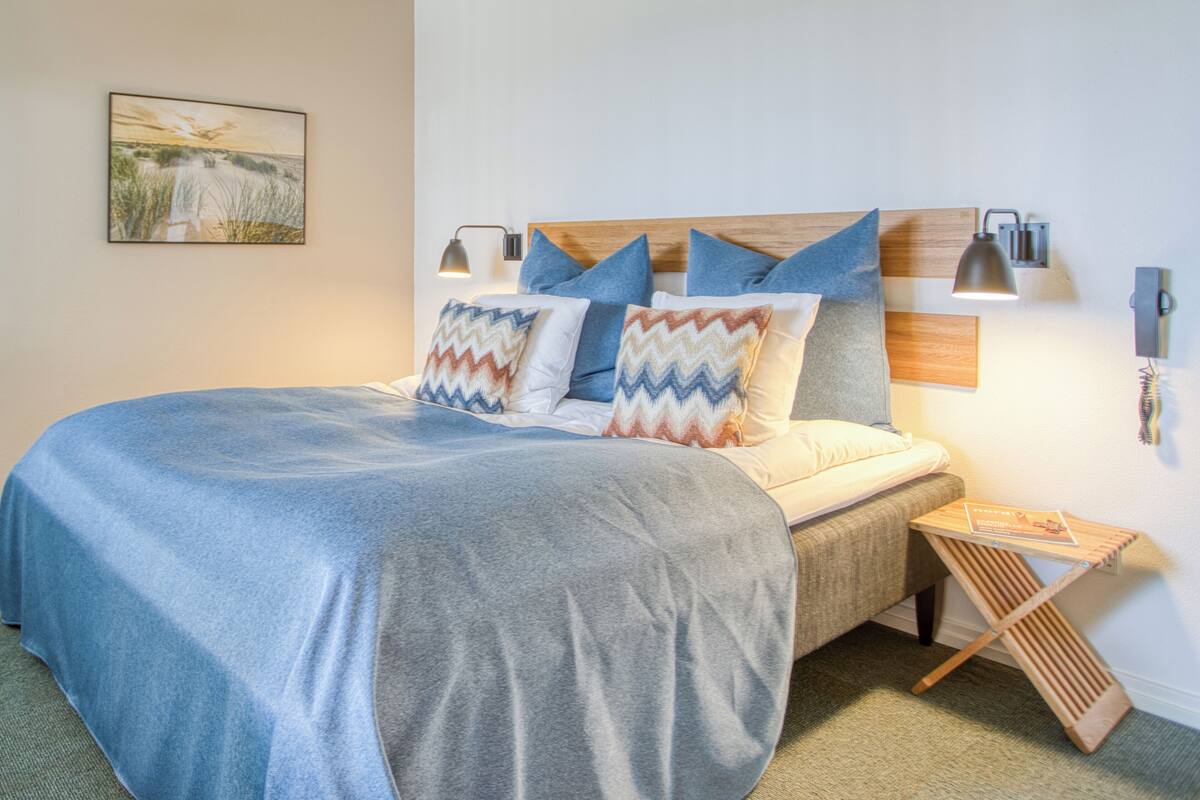 A neatly made bed with a blue blanket and patterned pillows, a bedside table with a book, and a wall-mounted lamp.