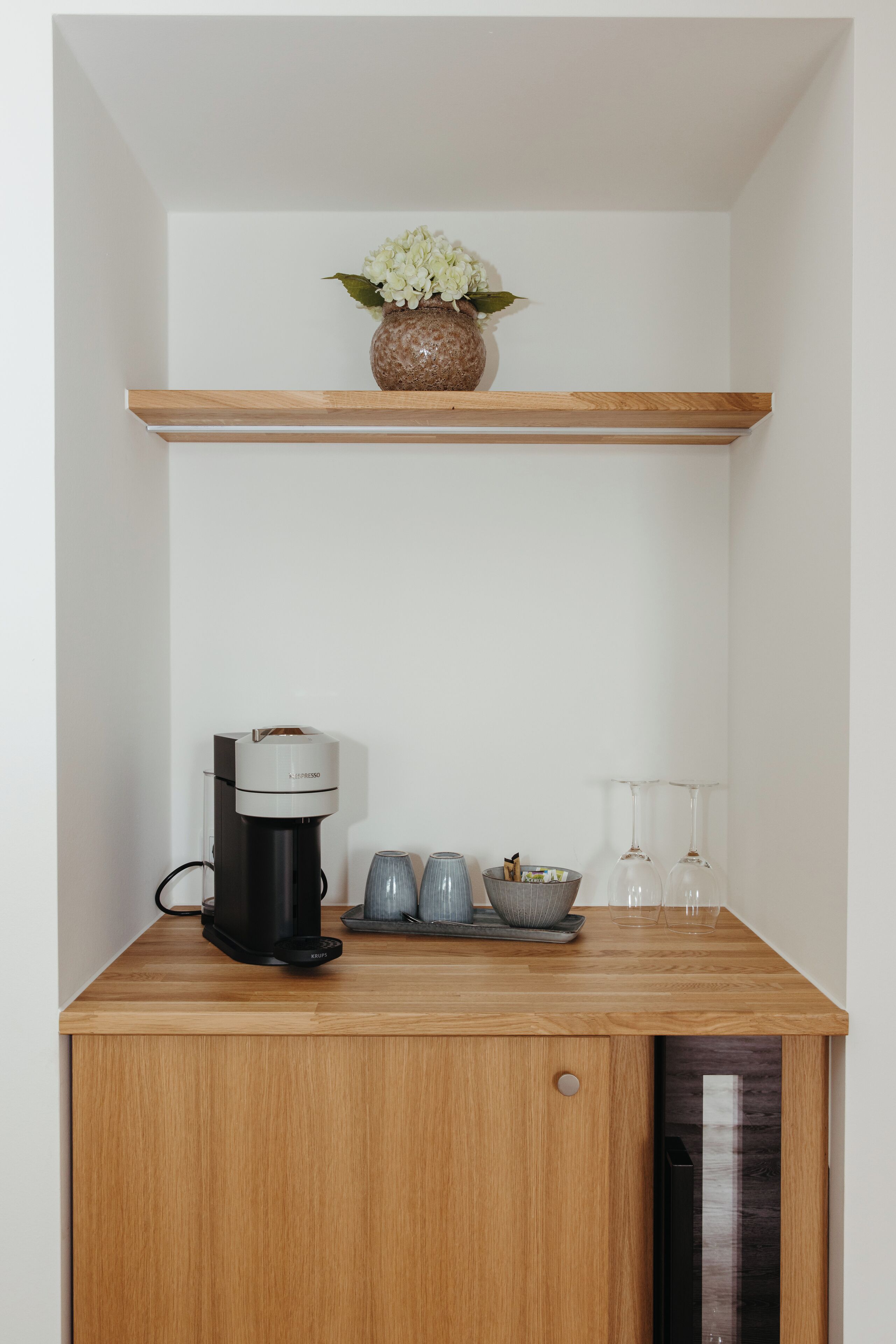 A kitchen with a coffee maker, two glasses, and a vase with flowers on a shelf.