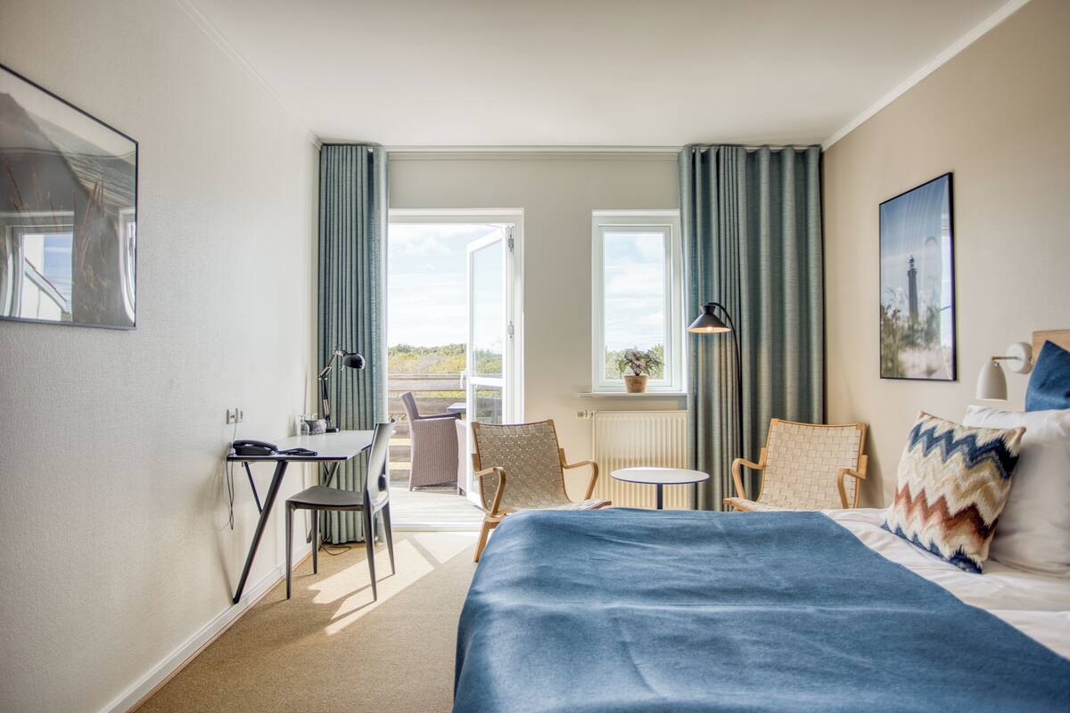A hotel room with a bed, desk, chairs, a small table, and a balcony with a view.