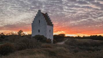 Una torre blanca solitaria con un techo empinado, rodeada de pasto seco y arbustos, bajo un cielo al atardecer.