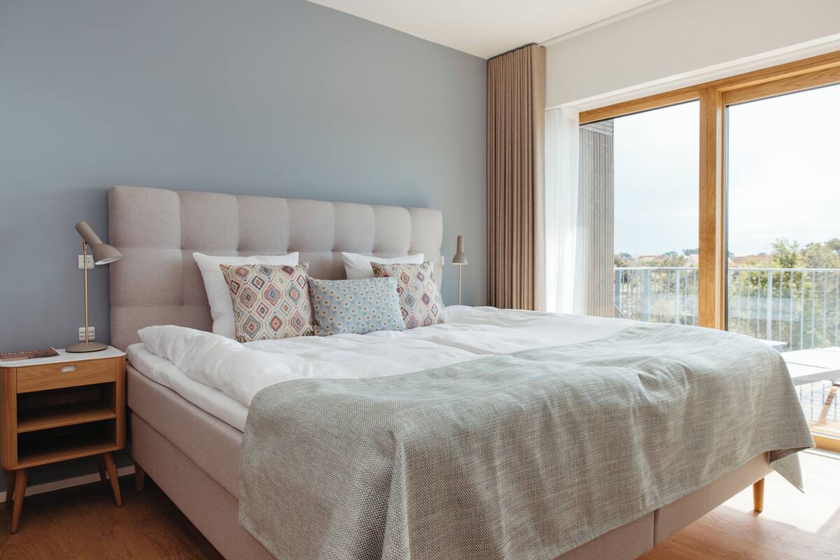 A modern bedroom with a large bed, two bedside tables, and a view of the outdoors through a large window.