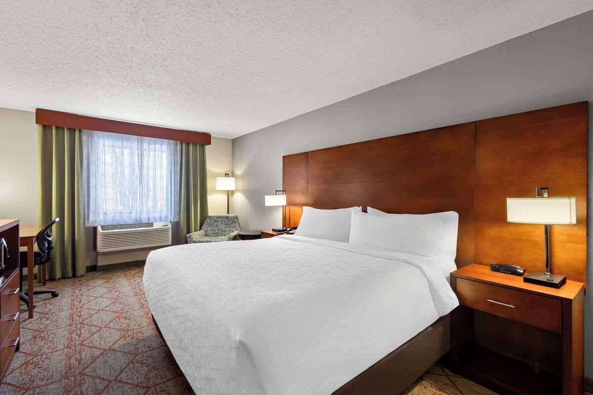 Ein Hotelzimmer mit einem großen Bett, zwei Nachttischen mit Lampen, einem Schreibtisch mit Stuhl und einem Fenster mit Vorhängen.