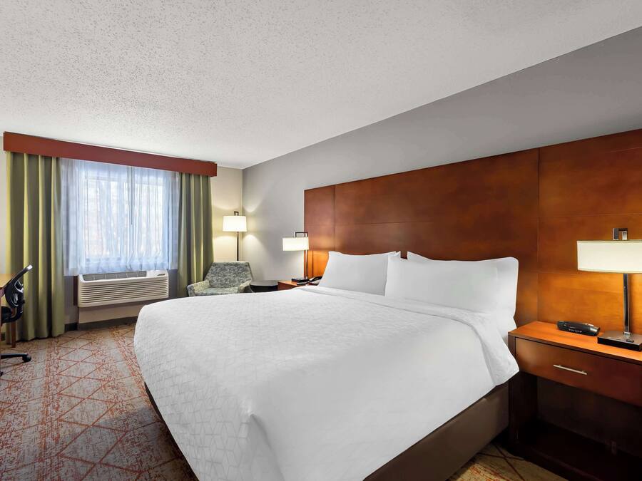 A hotel room with a large bed, two bedside tables with lamps, a desk with a chair, and a window with curtains.