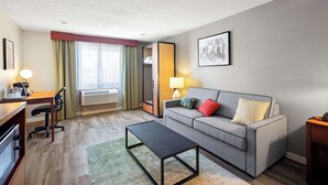 Quarto de hotel moderno com sofá cinza, escrivaninha de madeira, mesa de centro preta e tapete verde.