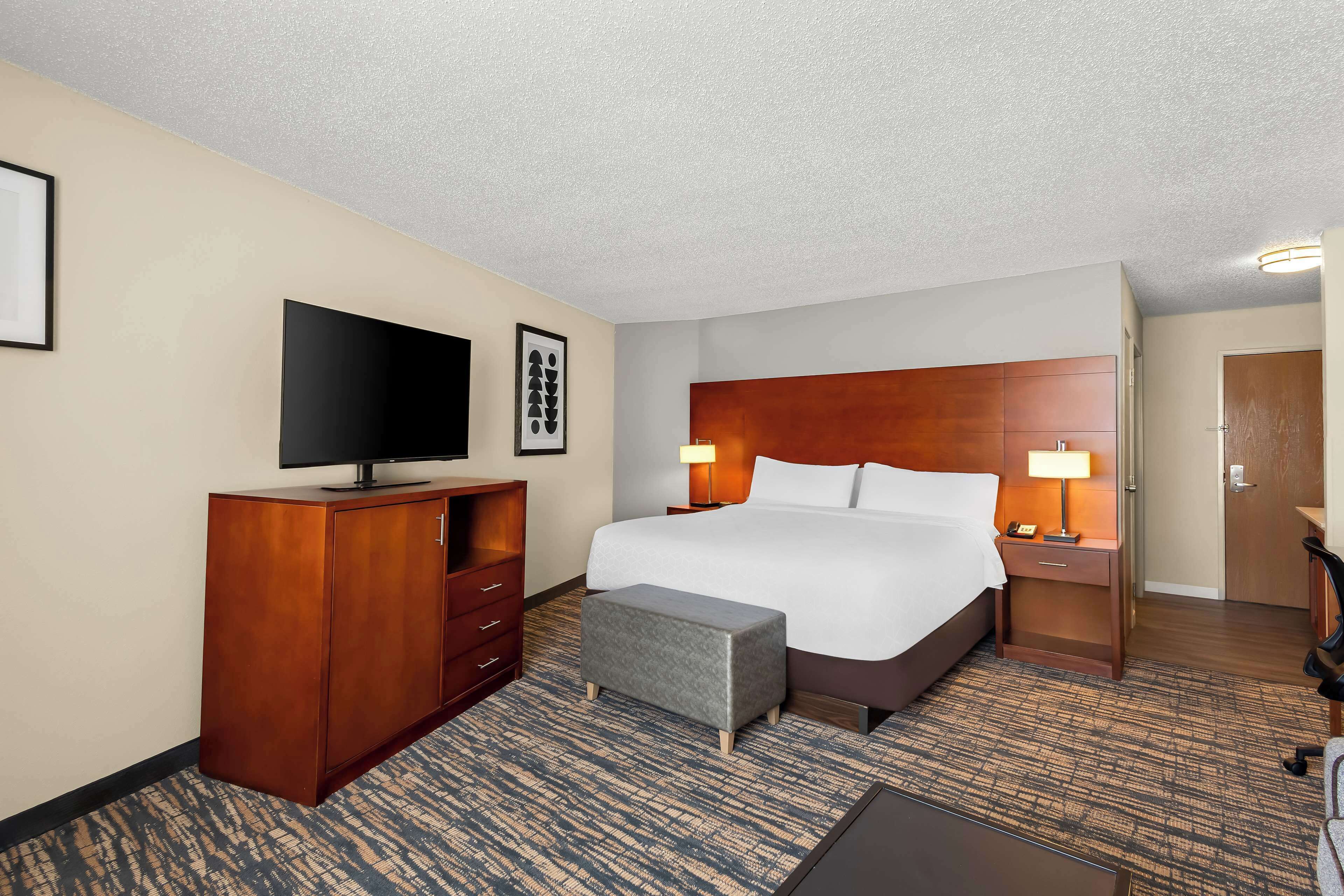 A hotel room with a large bed, a flat-screen TV, a wooden dresser, a bench, and a desk with a lamp.