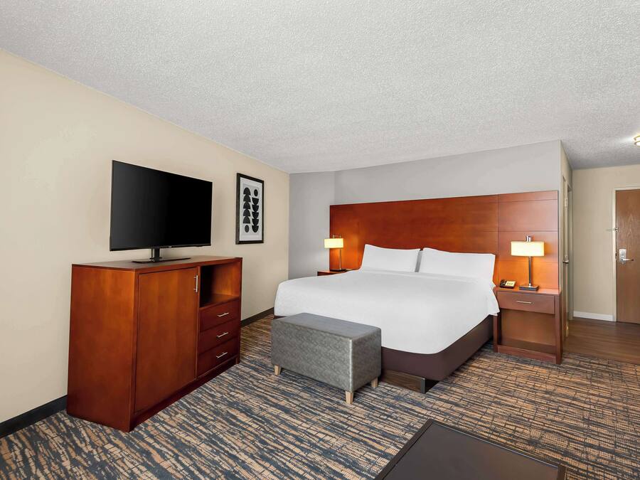 A hotel room with a large bed, a flat-screen TV, a wooden dresser, a bench, and a desk with a lamp.