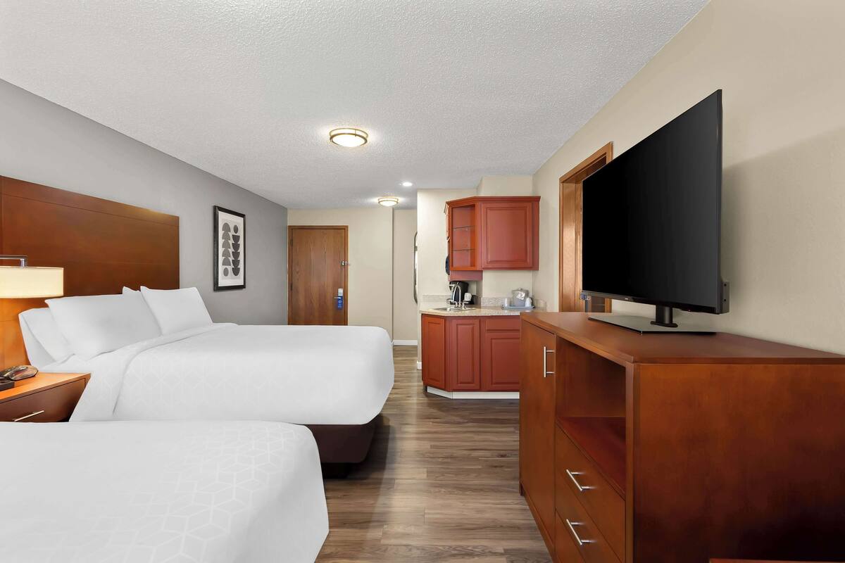 Ein Hotelzimmer mit zwei Betten, einem Fernseher, einem Schreibtisch und einer Küchenzeile.