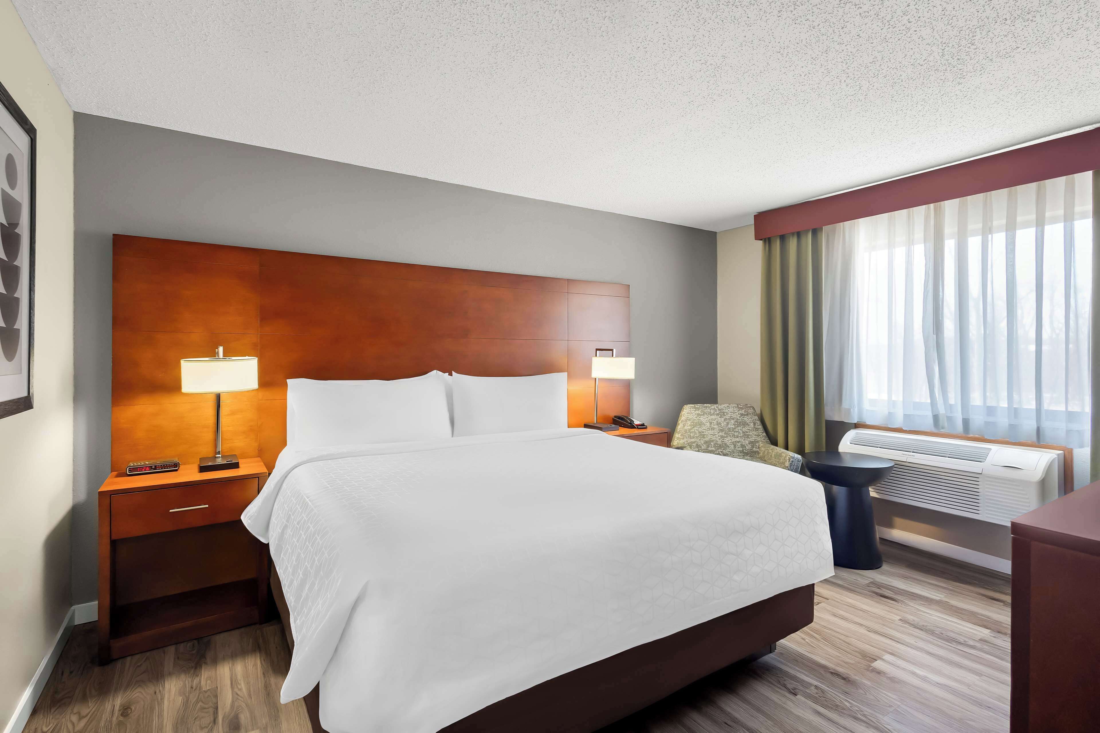 Una habitación de hotel con una cama grande, dos mesitas de noche con lámparas, una silla, un escritorio y un ventanal con cortinas.