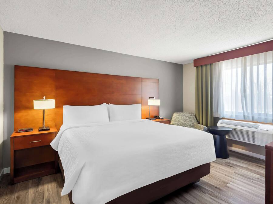 Una habitación de hotel con una cama grande, dos mesitas de noche con lámparas, una silla, un escritorio y un ventanal con cortinas.