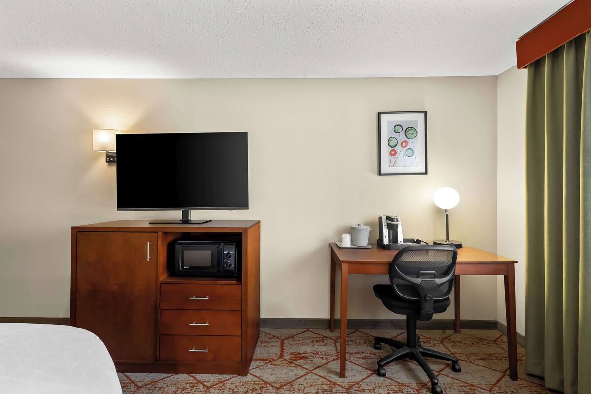 Ein Hotelzimmer mit einem Bett, einem hölzernen Schreibtisch mit Stuhl, einem Fernseher, einer Mikrowelle, einem gerahmten Bild an der Wand und einer Lampe.