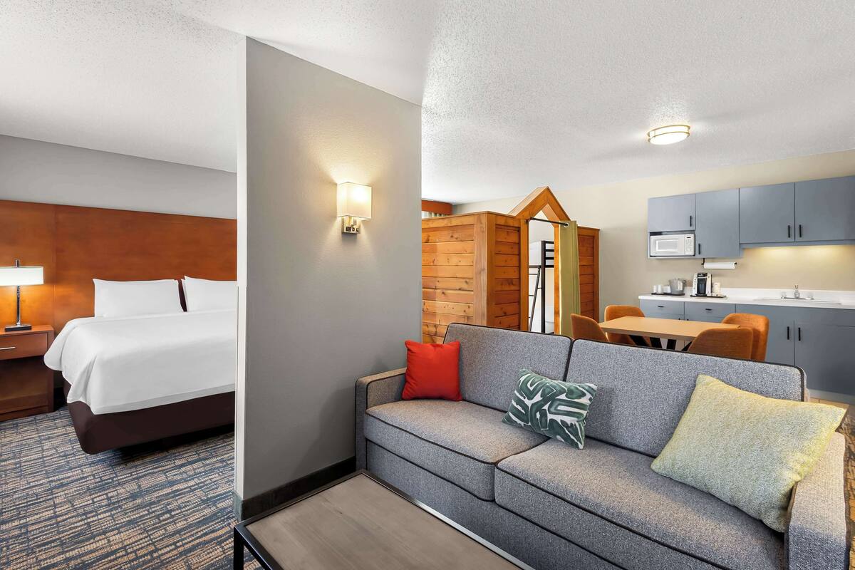 Ein Hotelzimmer mit Bett, Sofa, Holzwand, Küchenzeile mit Schränken und Mikrowelle sowie einer Leuchte.