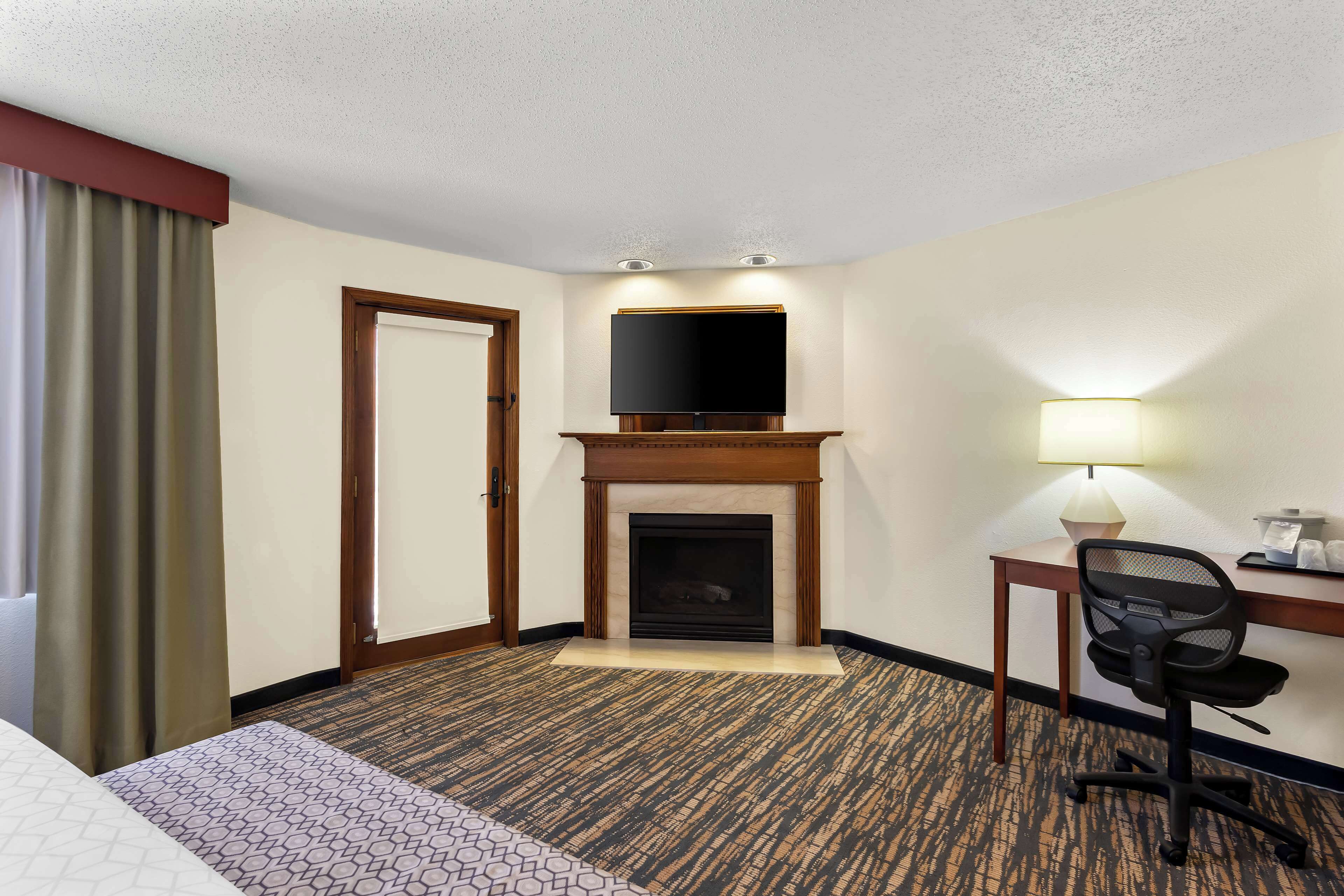 A hotel room with a bed, a desk with a chair, a television mounted above a fireplace, and a lamp on the desk.