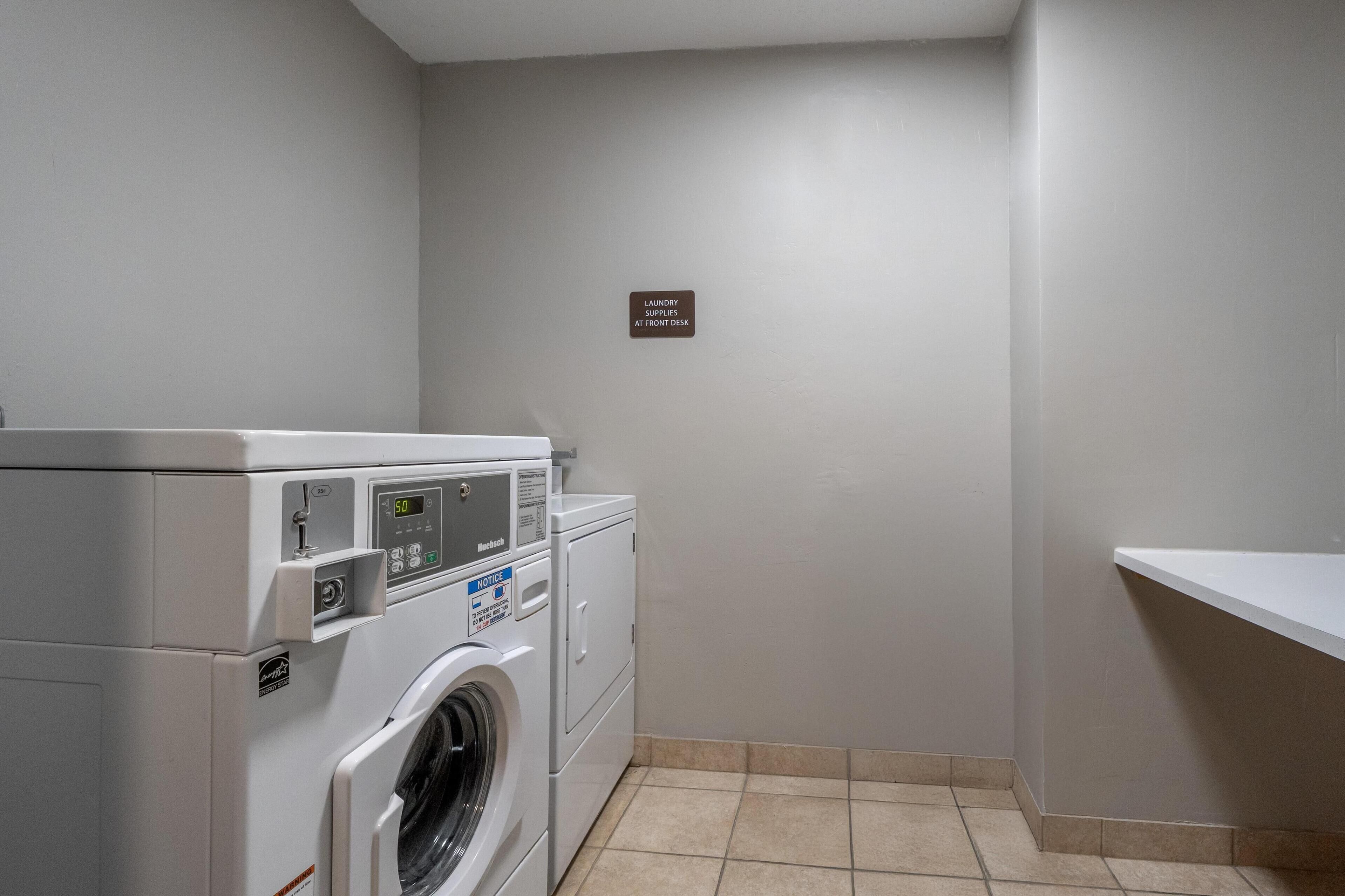 A laundry room with a front-loading washing machine and dryer, a sign indicating laundry supplies at the front desk, and a tiled floor.