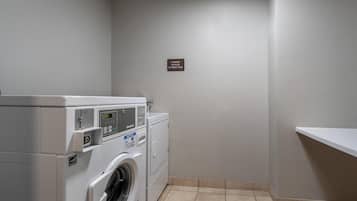A laundry room with a front-loading washing machine and dryer, a sign indicating laundry supplies at the front desk, and a tiled floor.