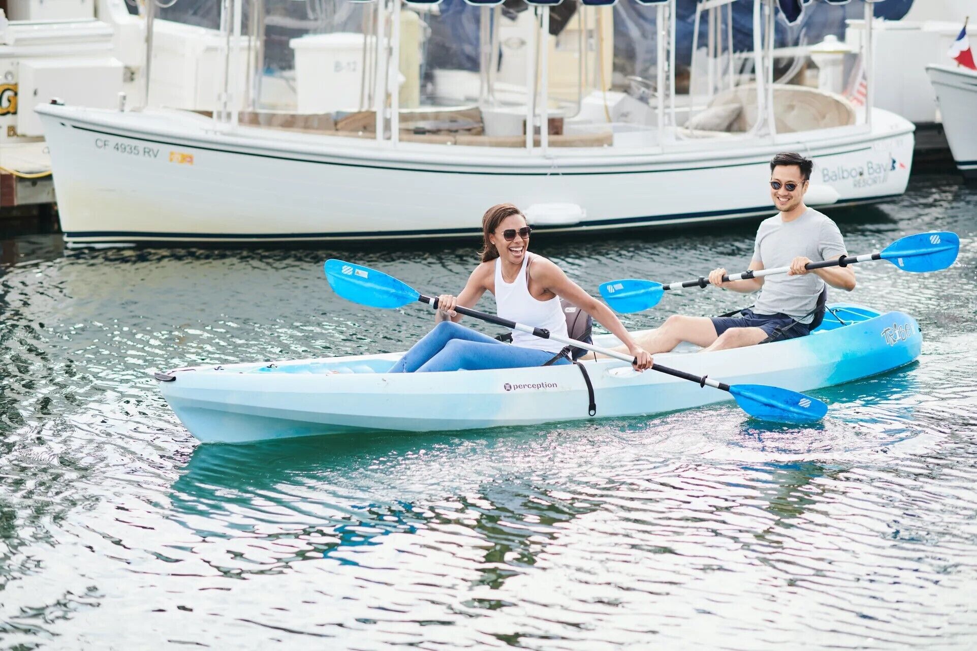 Two people kayaking on a calm body of water with boats in the background.