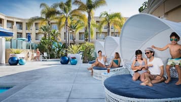 A poolside area with wicker seating and a covered area, palm trees, and a building with balconies in the background.