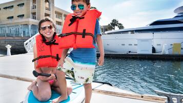 Two people on a boat with life jackets, a yacht docked nearby, and a building in the background.