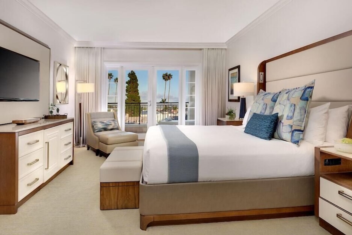 A hotel room with a large bed, a TV, a desk, a chair, and a balcony with a view.