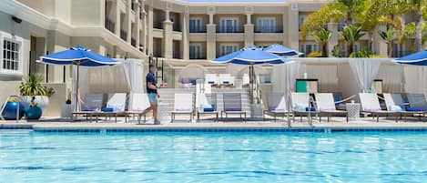 A pool area with lounge chairs and umbrellas, a building with balconies, and palm trees.