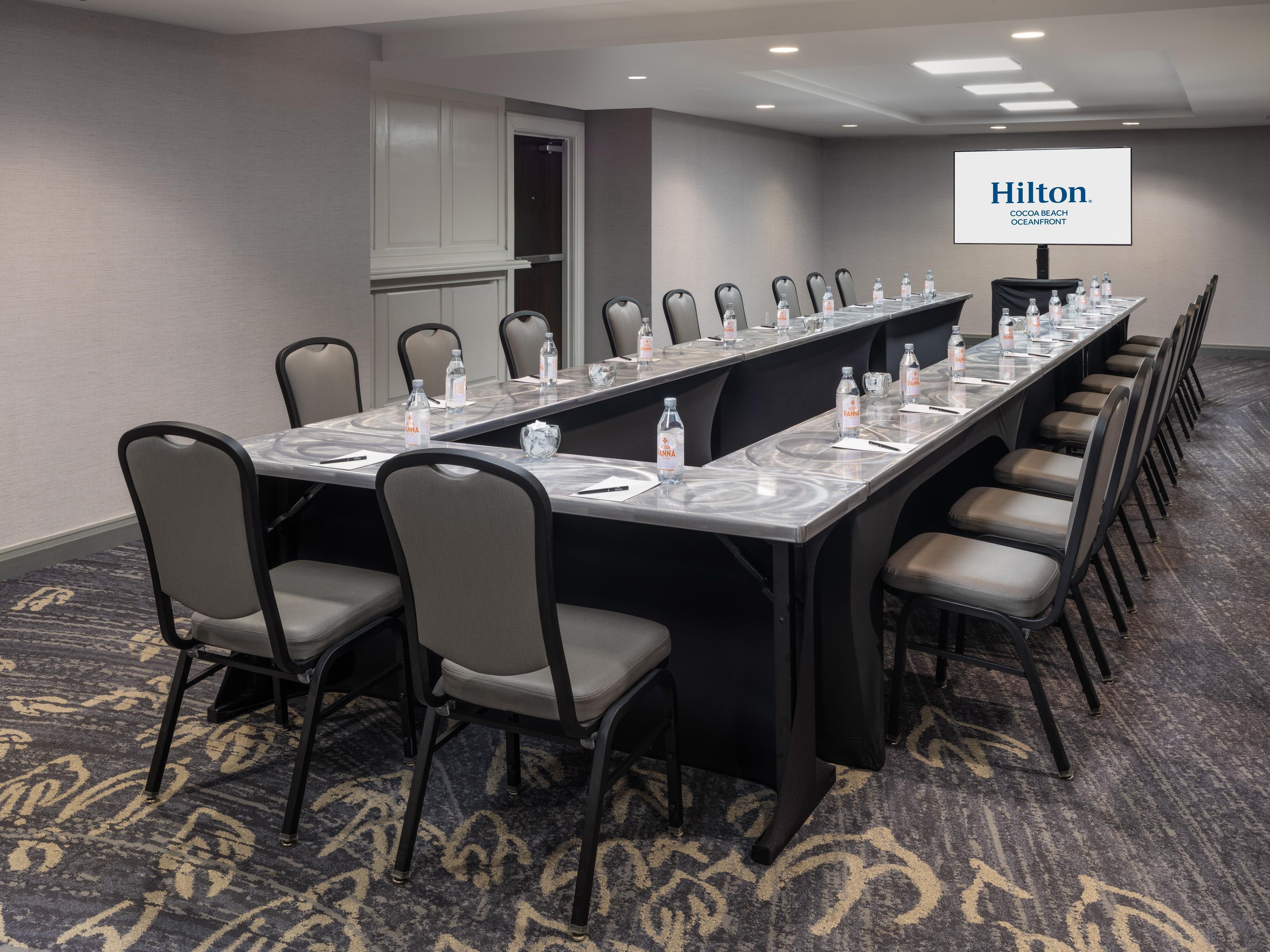 A conference room with a long table, chairs, water bottles, and a screen displaying 'Hilton Cocoa Beach Oceanfront'.