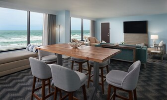 Habitación de hotel con un ventanal grande con vistas al océano, una mesa de comedor de madera con cuatro sillas, un sofá, un televisor y una pequeña mesita auxiliar.