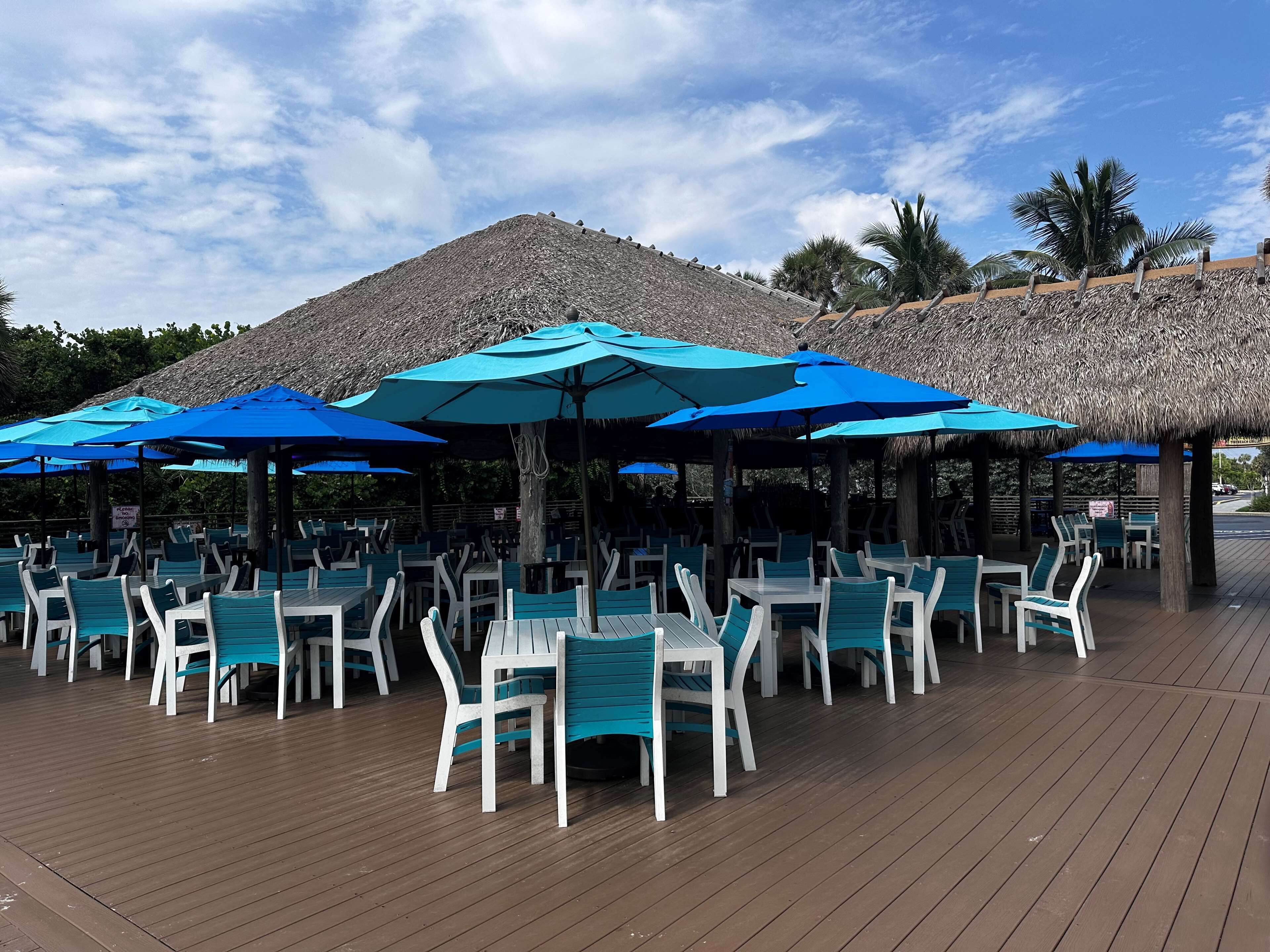 Un espace extérieur aménagé pour les repas, avec des tables et des chaises, des parasols bleus et une structure dotée d’un toit de chaume.
