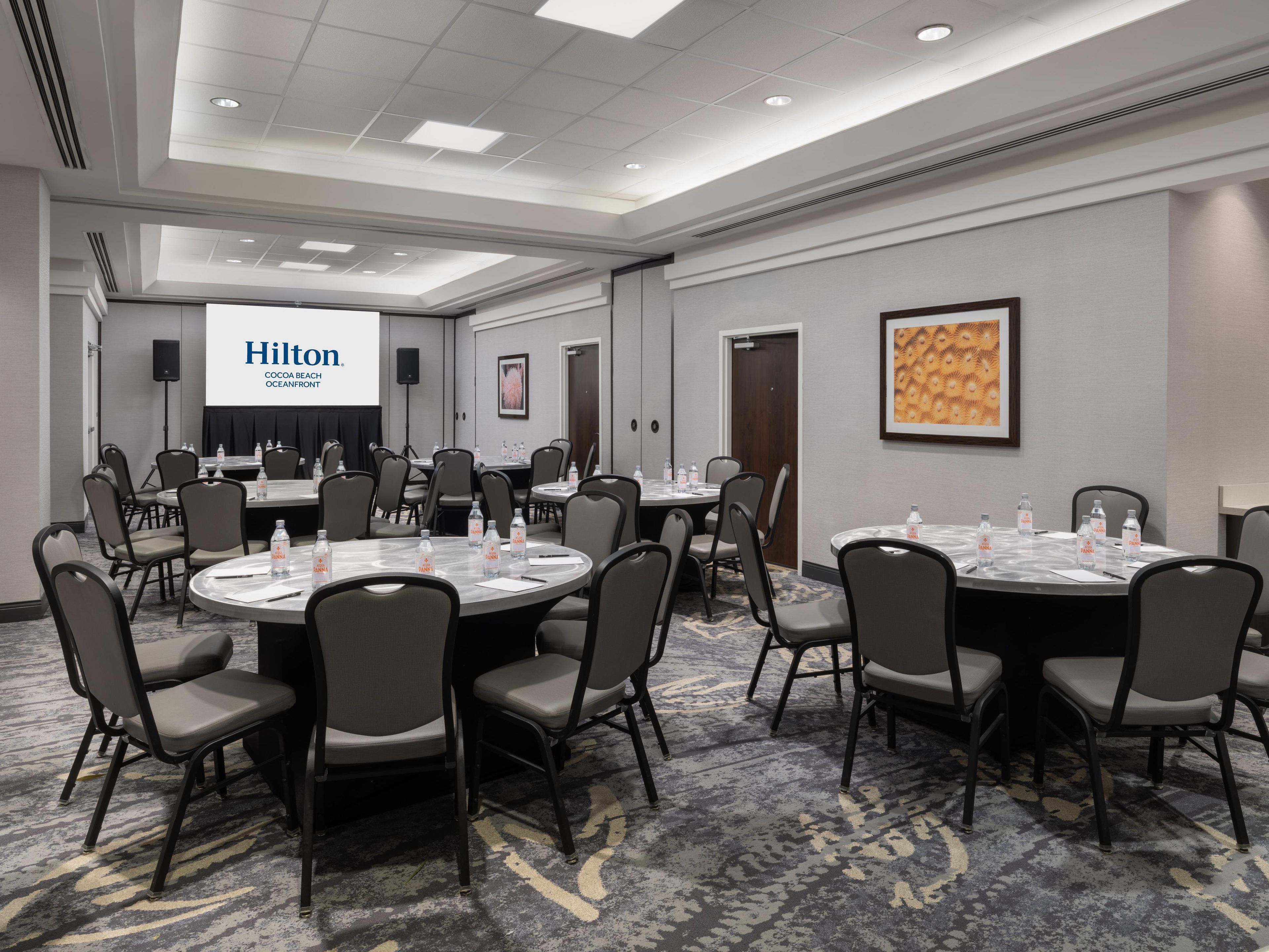 Une salle de conférence équipée de tables rondes, de chaises et d’un écran de projection affichant l’inscription « Hilton Cocoa Beach Oceanfront ».