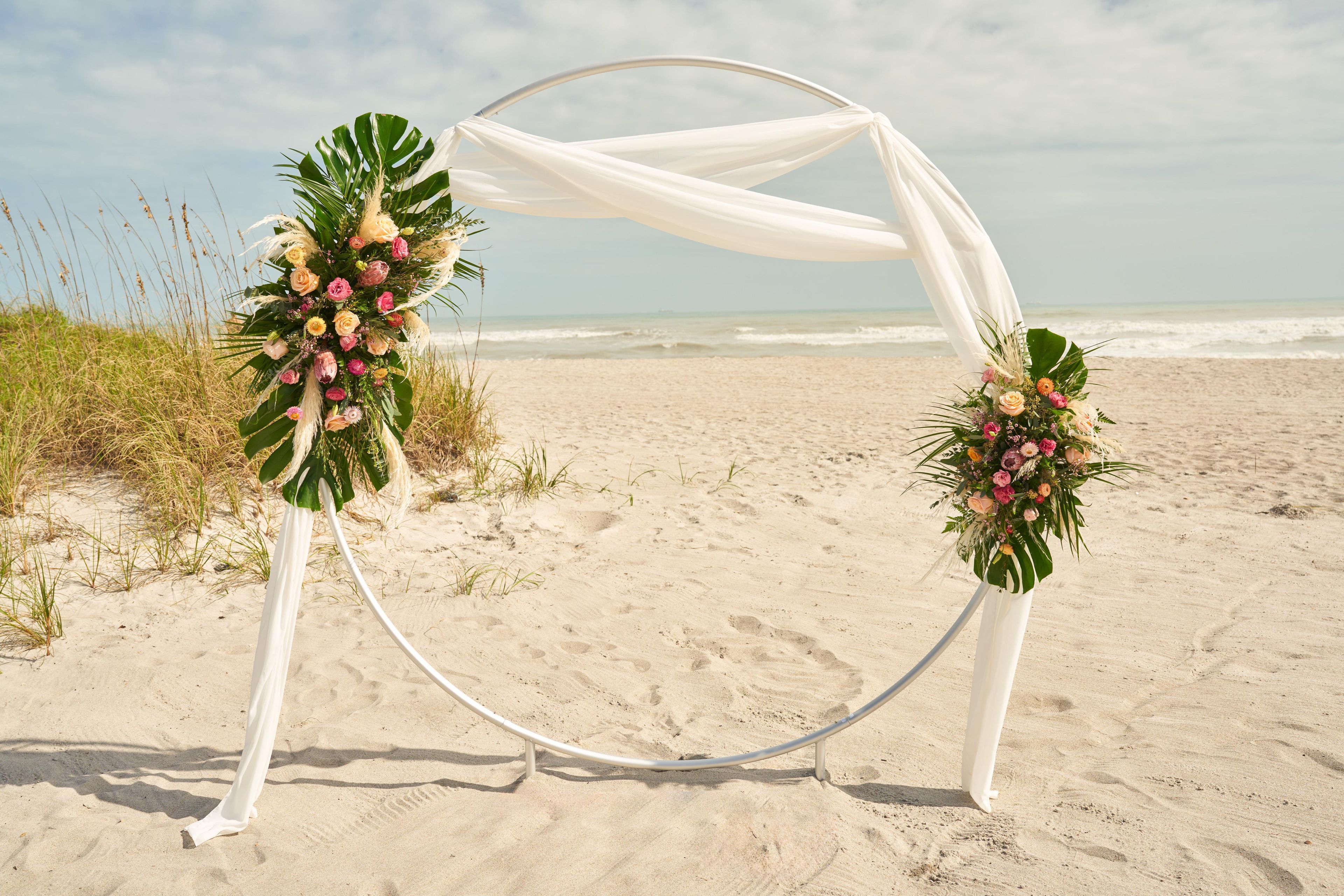 A beach wedding arch with white drapes and floral arrangements.