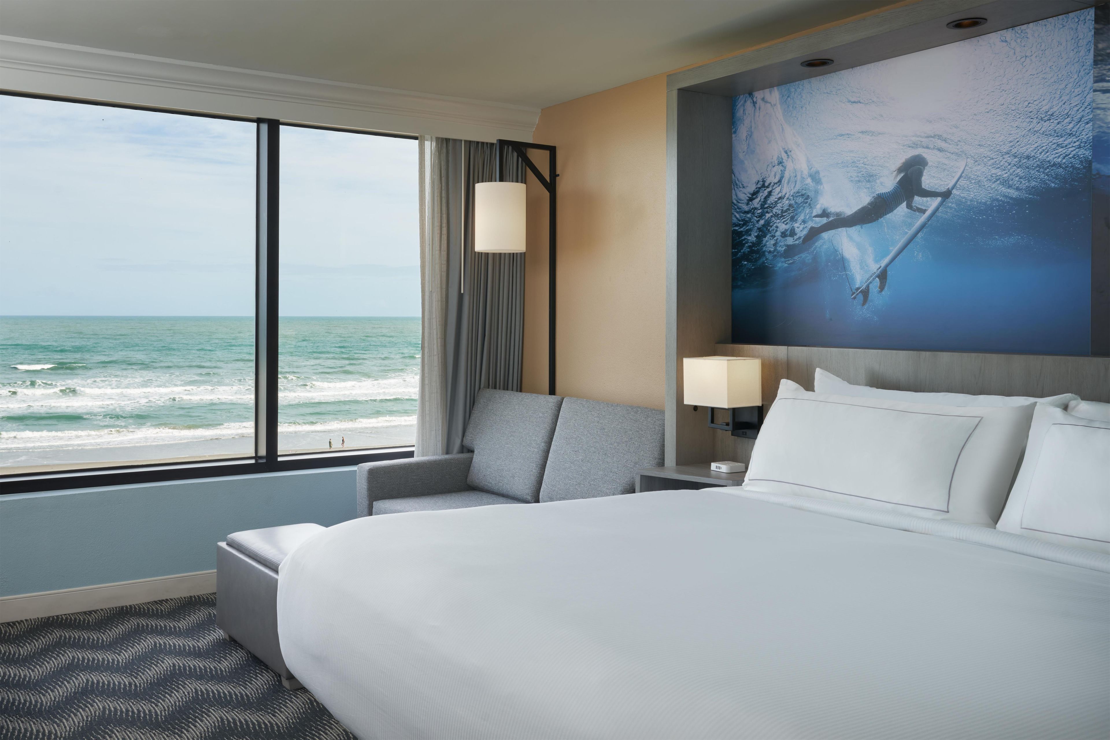 A hotel room with a large bed, a seating area, a view of the ocean, and a painting of a surfer.