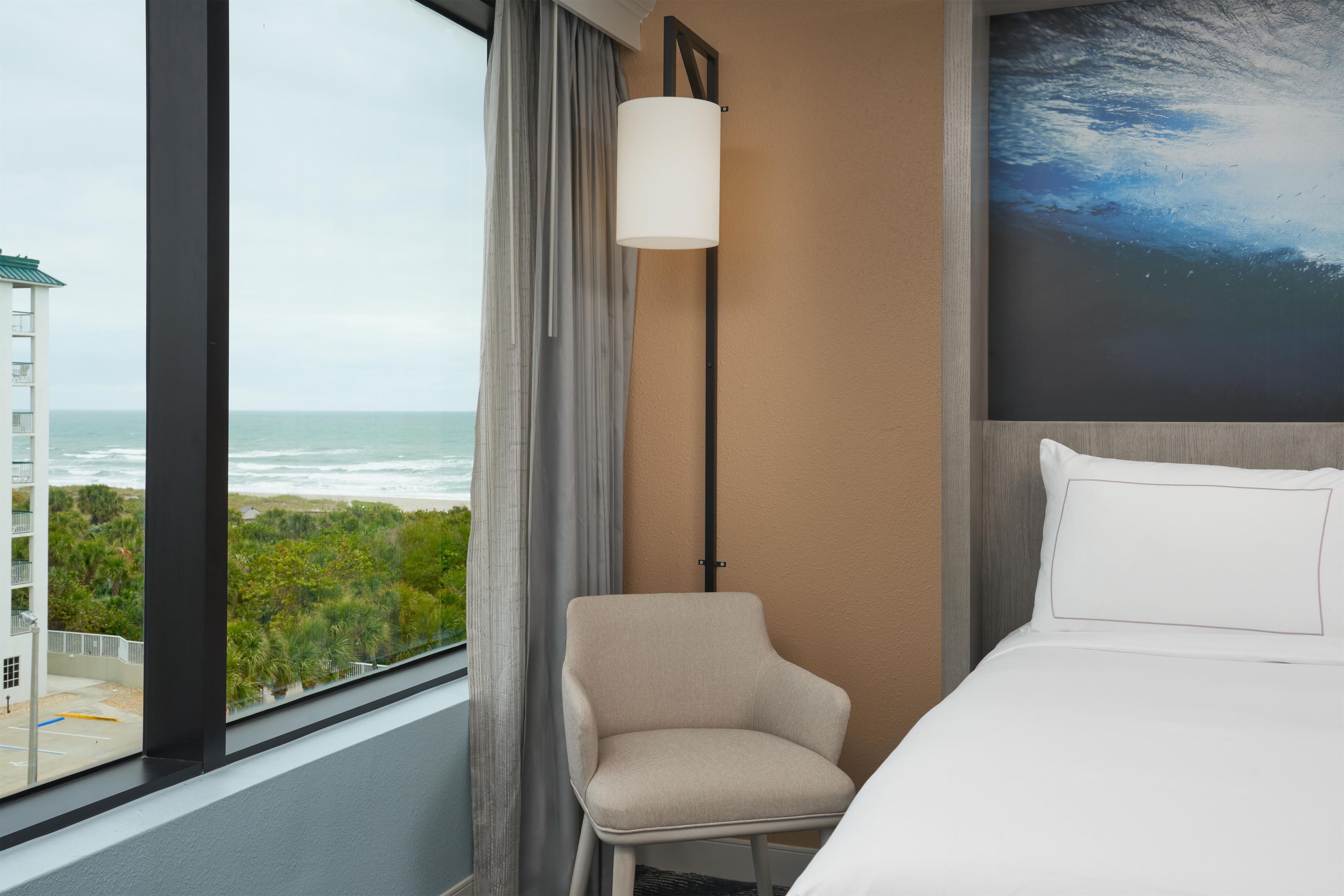 A hotel room with a bed, a chair, a view of the ocean, and a painting of the sky.