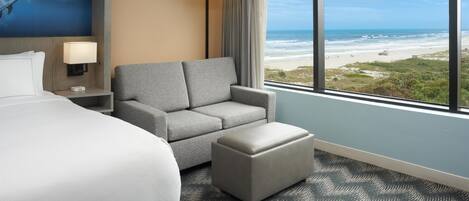 A hotel room with a large bed, a grey armchair, a small ottoman, a nightstand with a lamp, and a view of the beach.