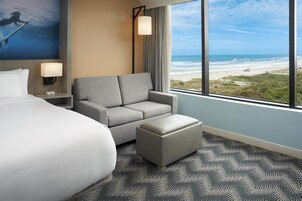 Habitación de hotel con una cama grande, un sillón gris, un pequeño reposapiés, una mesita de noche con lámpara y vista a la playa.