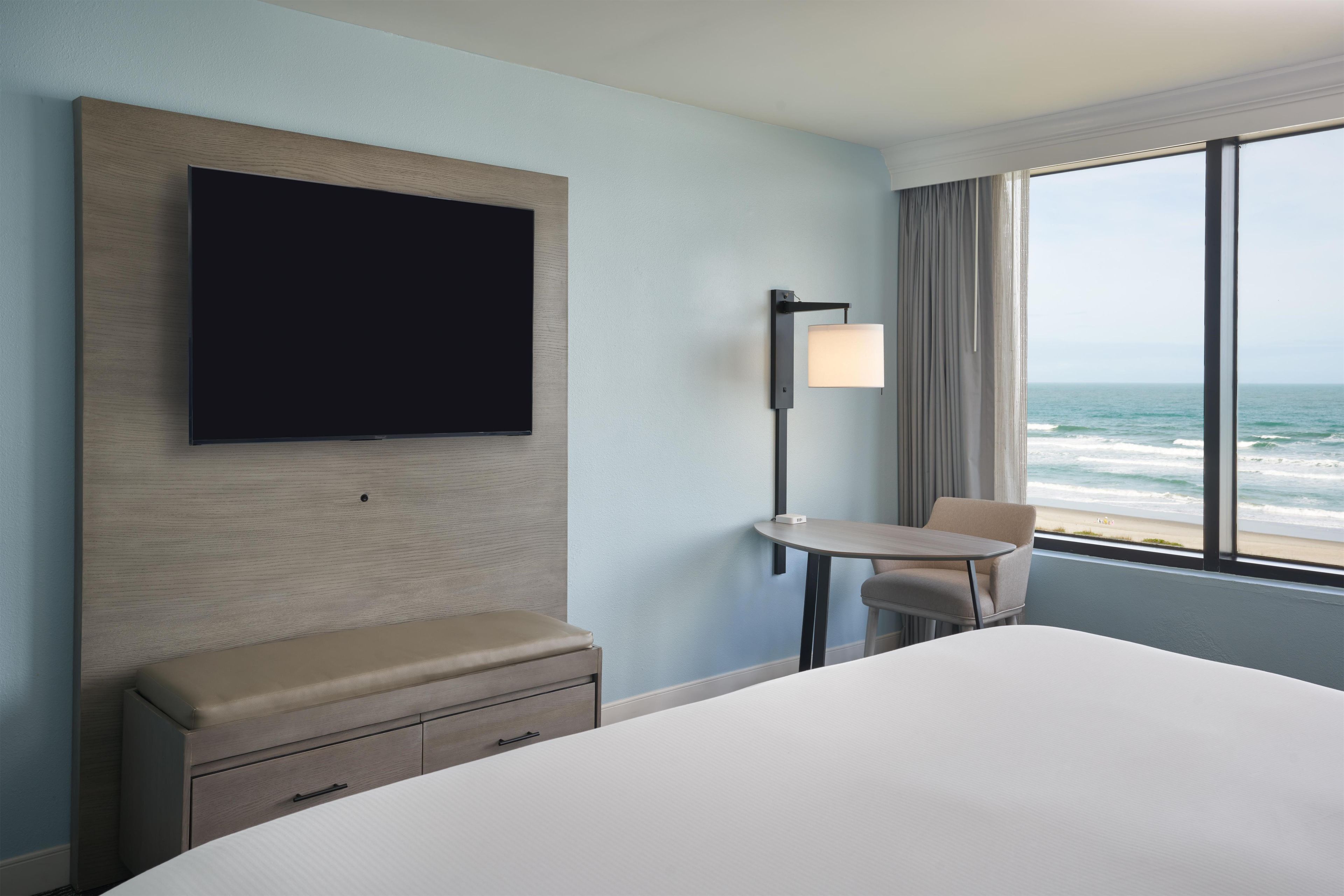 A hotel room with a bed, a TV mounted on the wall, a small table with a chair, and a view of the ocean through the window.