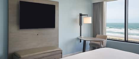 A hotel room with a bed, a TV mounted on the wall, a small table with a chair, and a view of the ocean through the window.