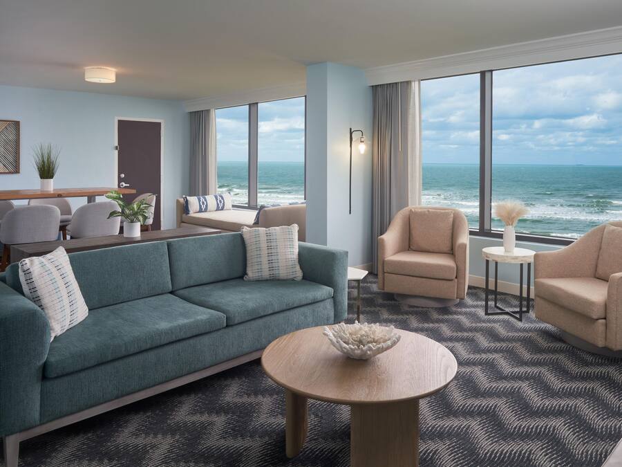 Habitación de hotel moderna con amplia vista al océano, un sofá, sillones, una mesa de comedor y una mesita auxiliar.