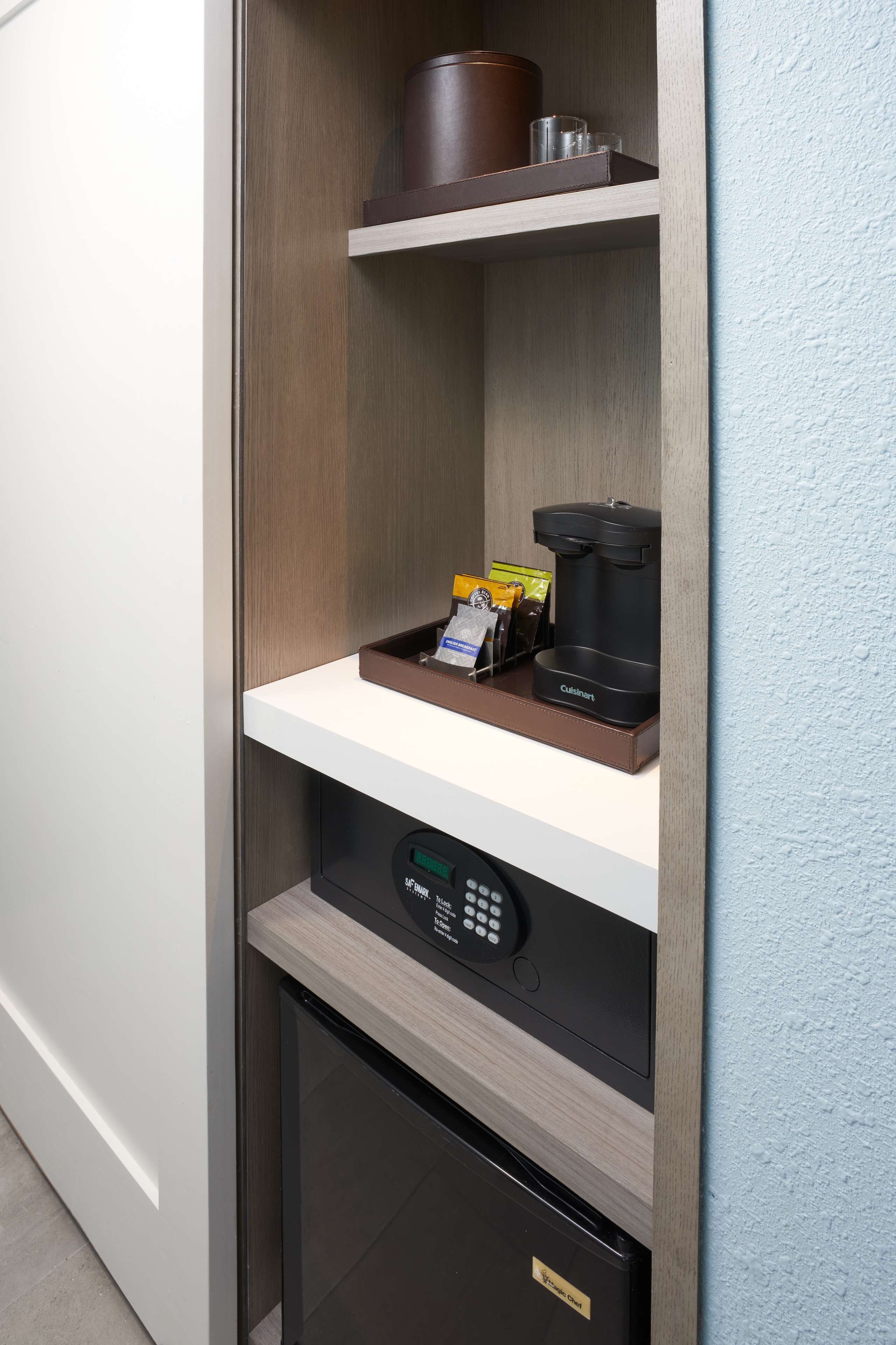 A kitchenette with a coffee maker, a small refrigerator, and a shelf with a brown container and a glass.