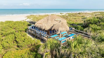 Un complexe hôtelier en bord de mer, doté d’une structure à toit de chaume, de plusieurs espaces de détente avec des chaises bleues et offrant une vue sur l’océan.