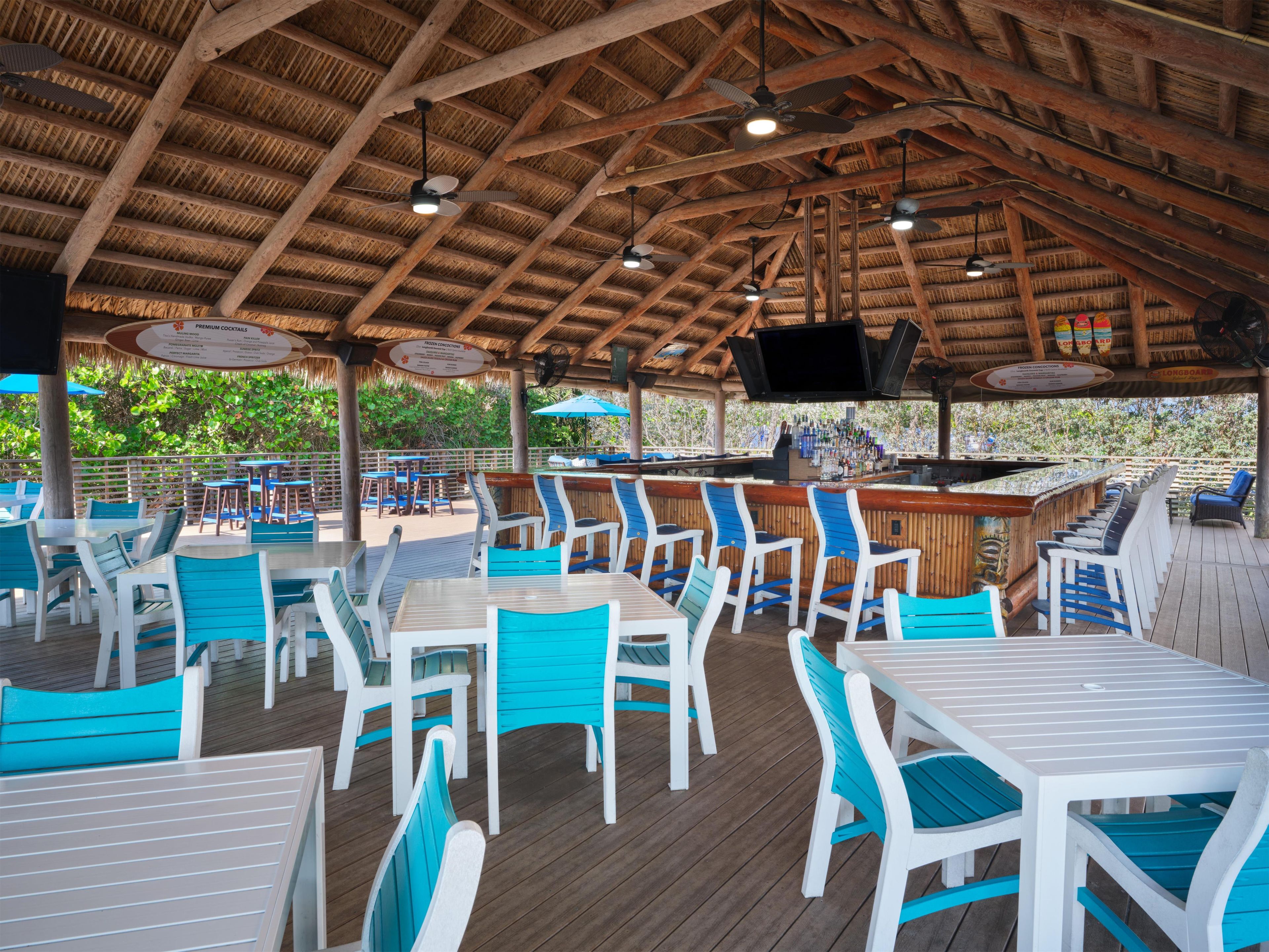 Un área de bar al aire libre con vigas de madera en el techo, varias mesas y sillas, y una barra con pantalla de televisión.