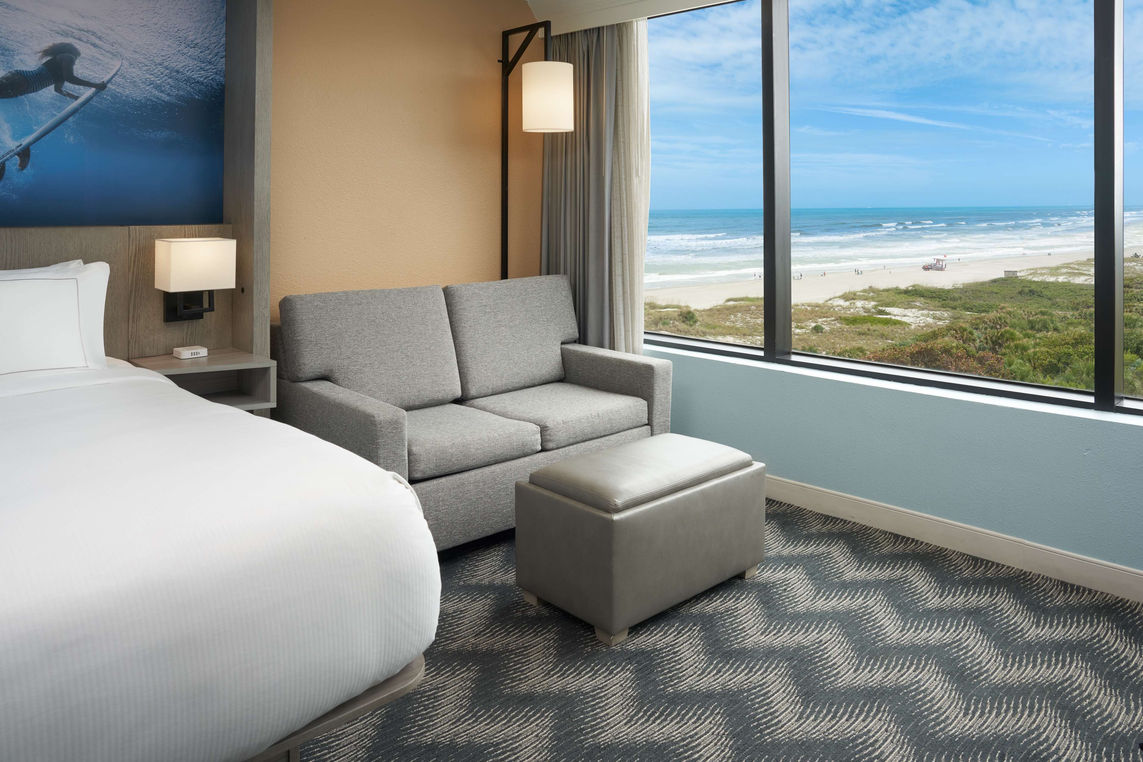 Ein Hotelzimmer mit einem Bett, einem grauen Sofa, einem Hocker, einem Nachttisch mit Lampe und einem großen Fenster mit Blick auf den Strand.