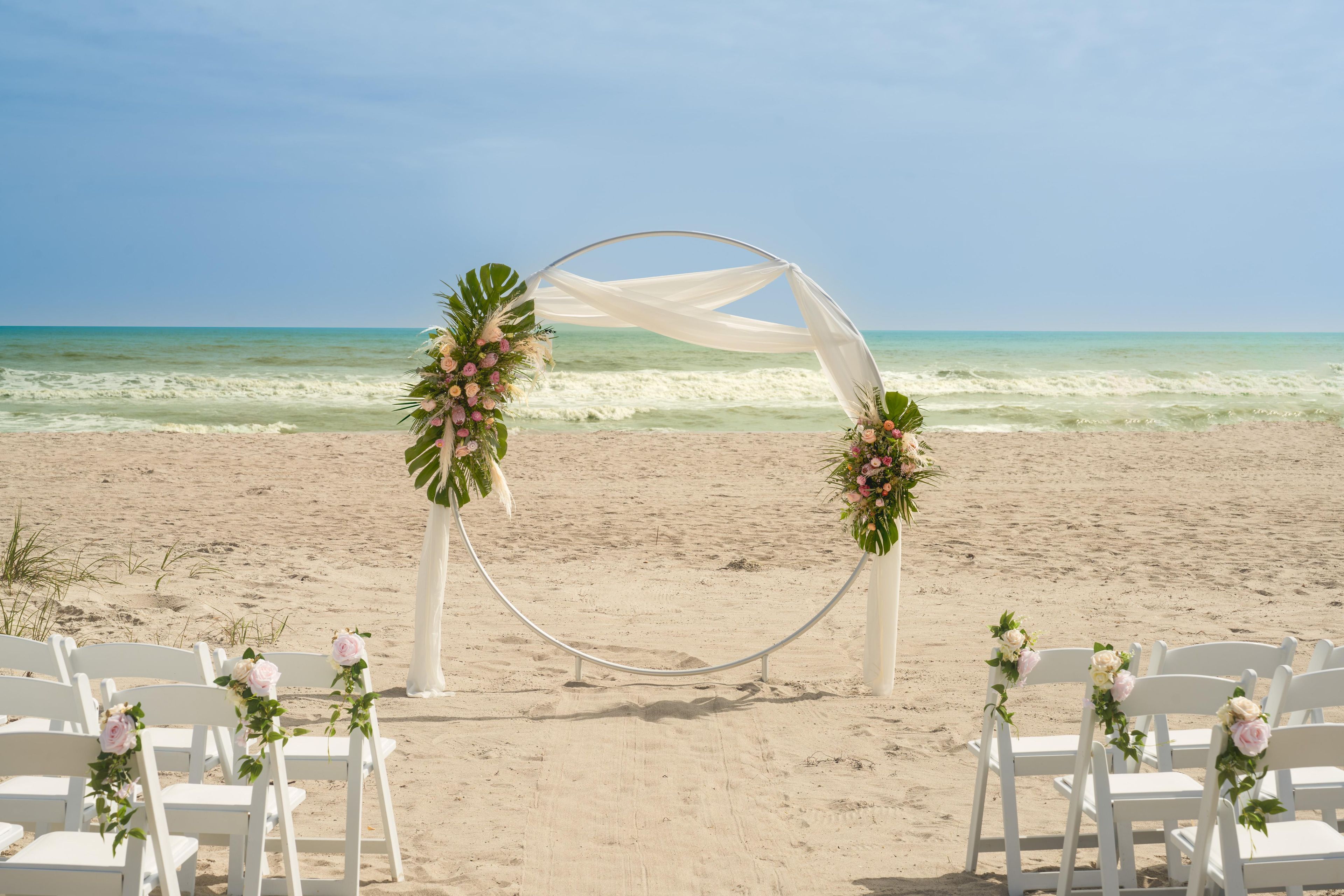 A beach wedding setup with a white arch and floral decorations, white chairs arranged in rows, and the ocean in the background.