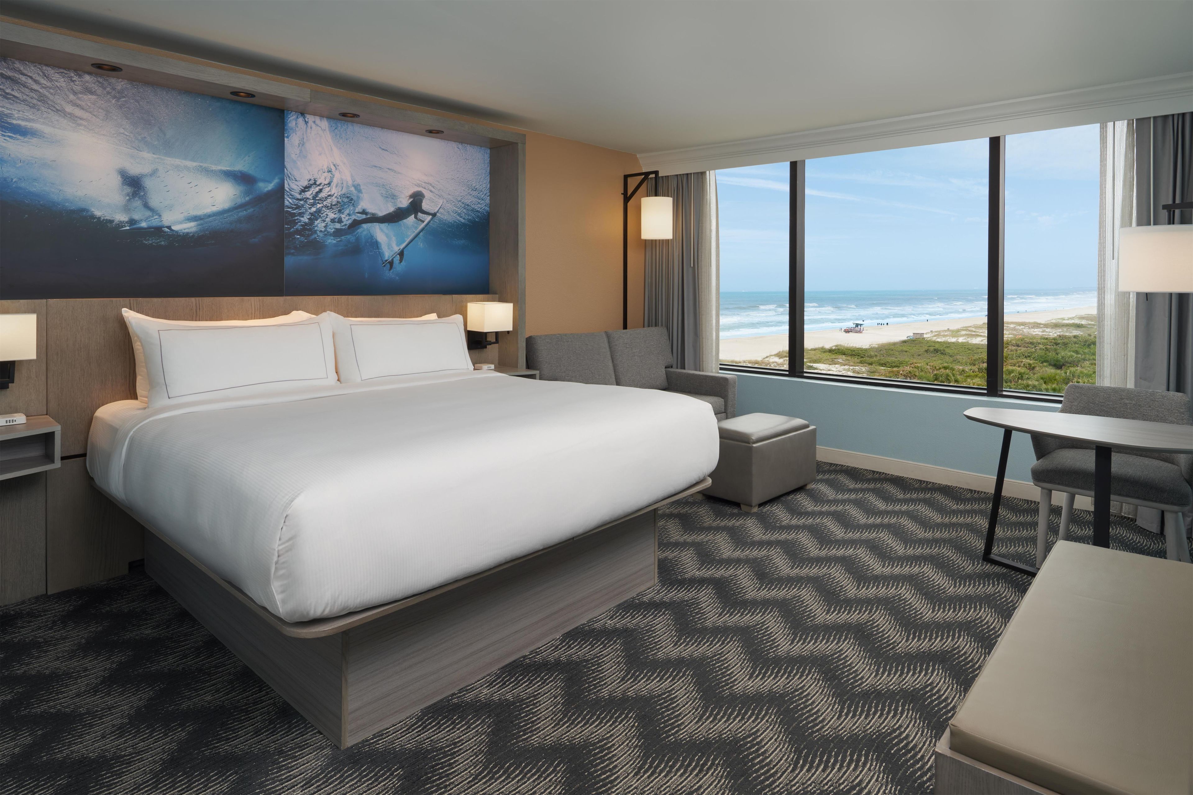 A hotel room with a large bed, a desk, a chair, and a view of the beach.