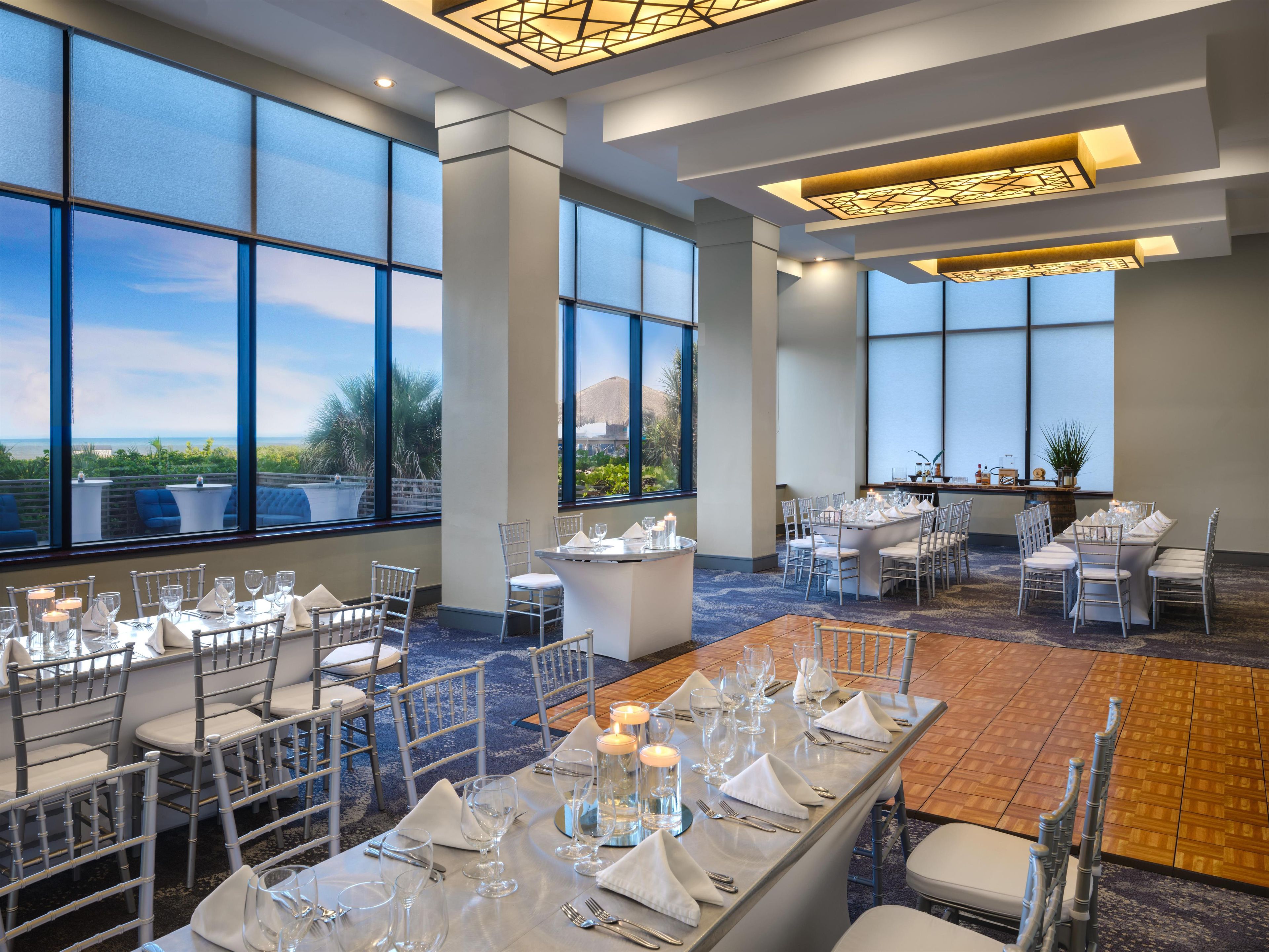 A spacious dining area with high ceilings, large windows, and a view of the outdoors. Tables are set with white tablecloths, silverware, and candles.