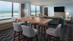 A hotel room with a wooden dining table, four chairs, a sofa, a TV, and a view of the ocean. - Hilton Cocoa Beach Oceanfront (Cocoa Beach)
