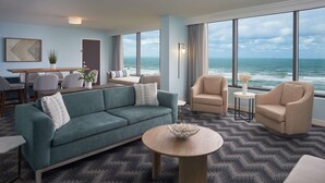 A modern hotel room with a large ocean view, a green sofa, two armchairs, a dining area, and a small side table with a decorative bowl. - Hilton Cocoa Beach Oceanfront (Cocoa Beach)