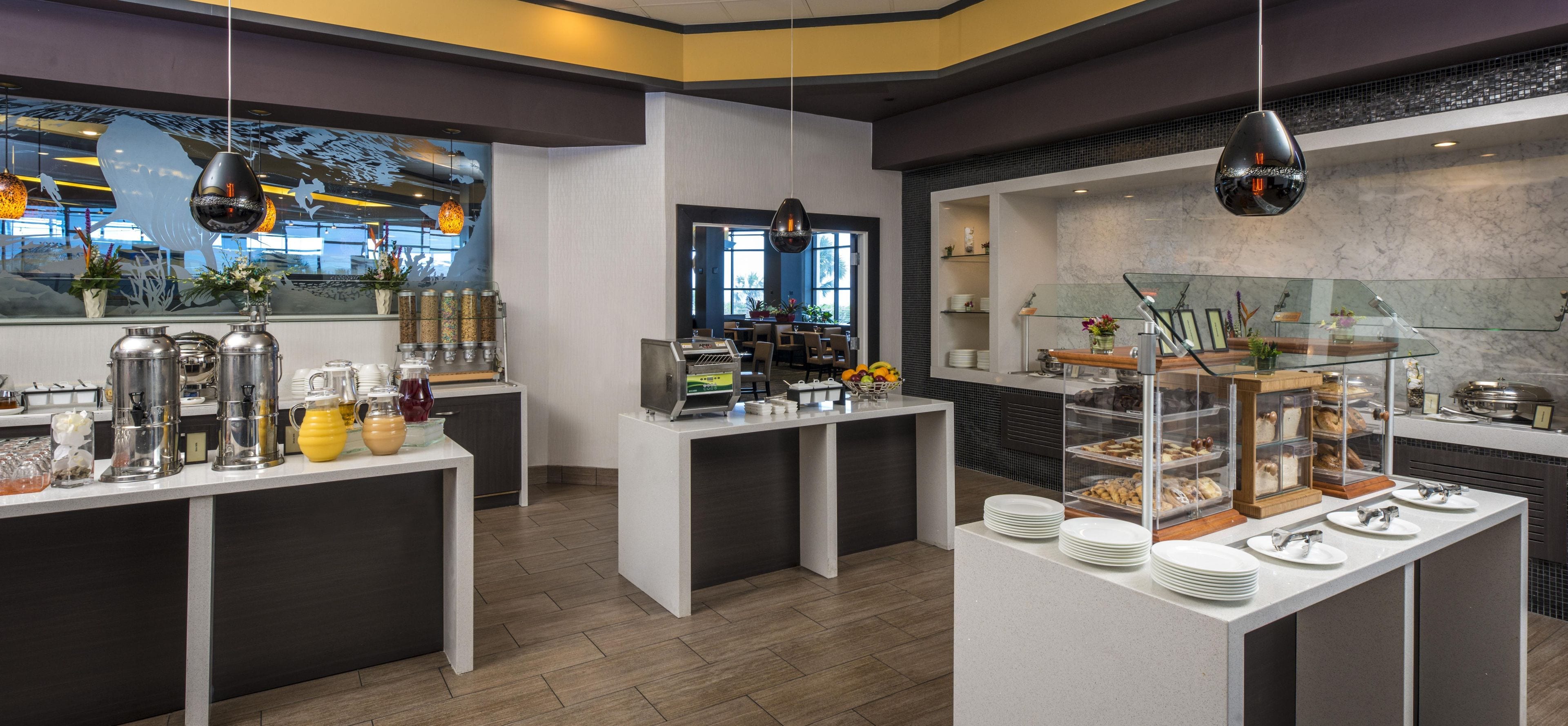 A modern breakfast area with self-service stations, including coffee dispensers, fruit, and pastries.