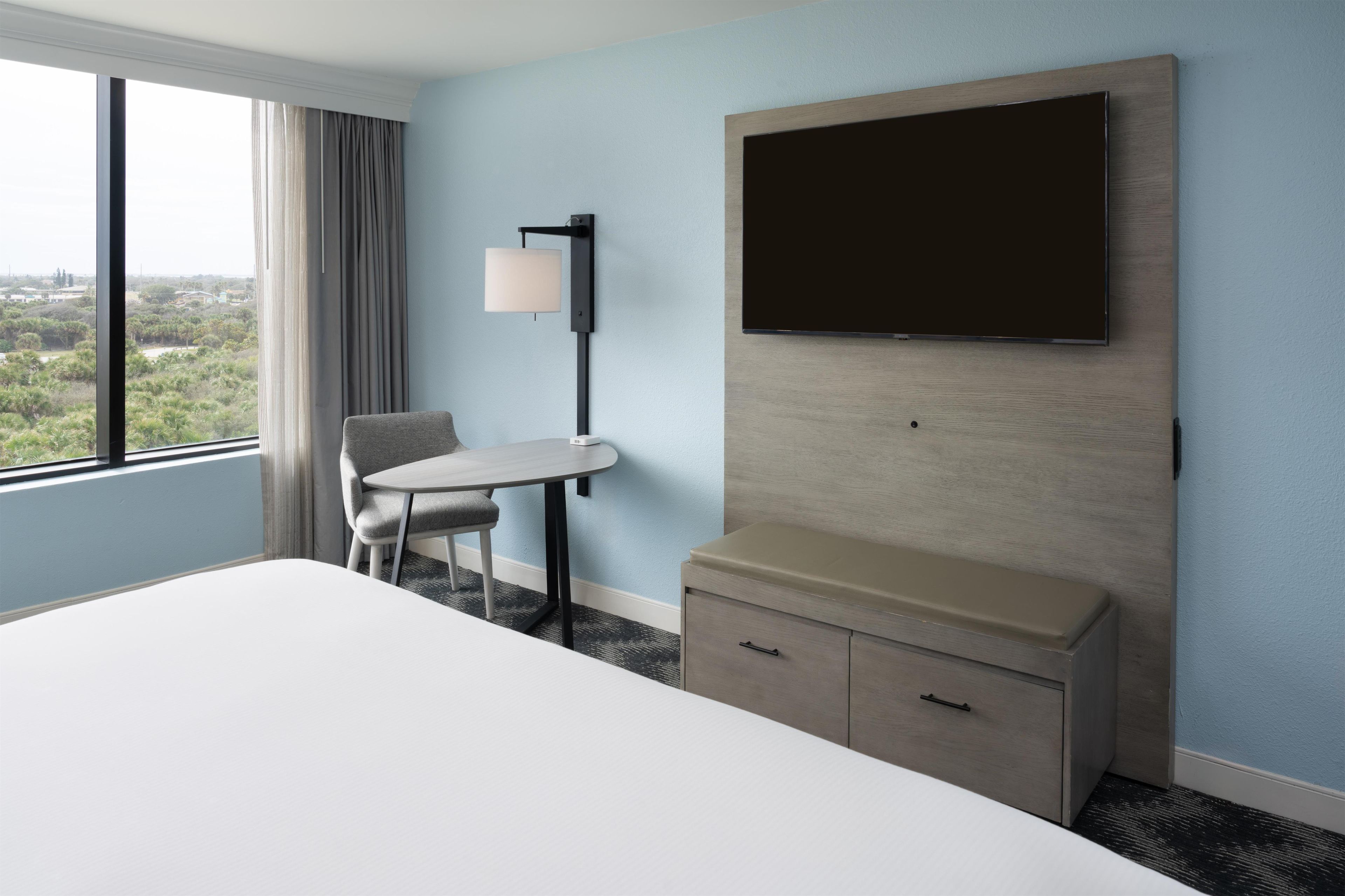 A hotel room with a bed, a desk, a chair, a TV mounted on the wall, and a window with curtains.