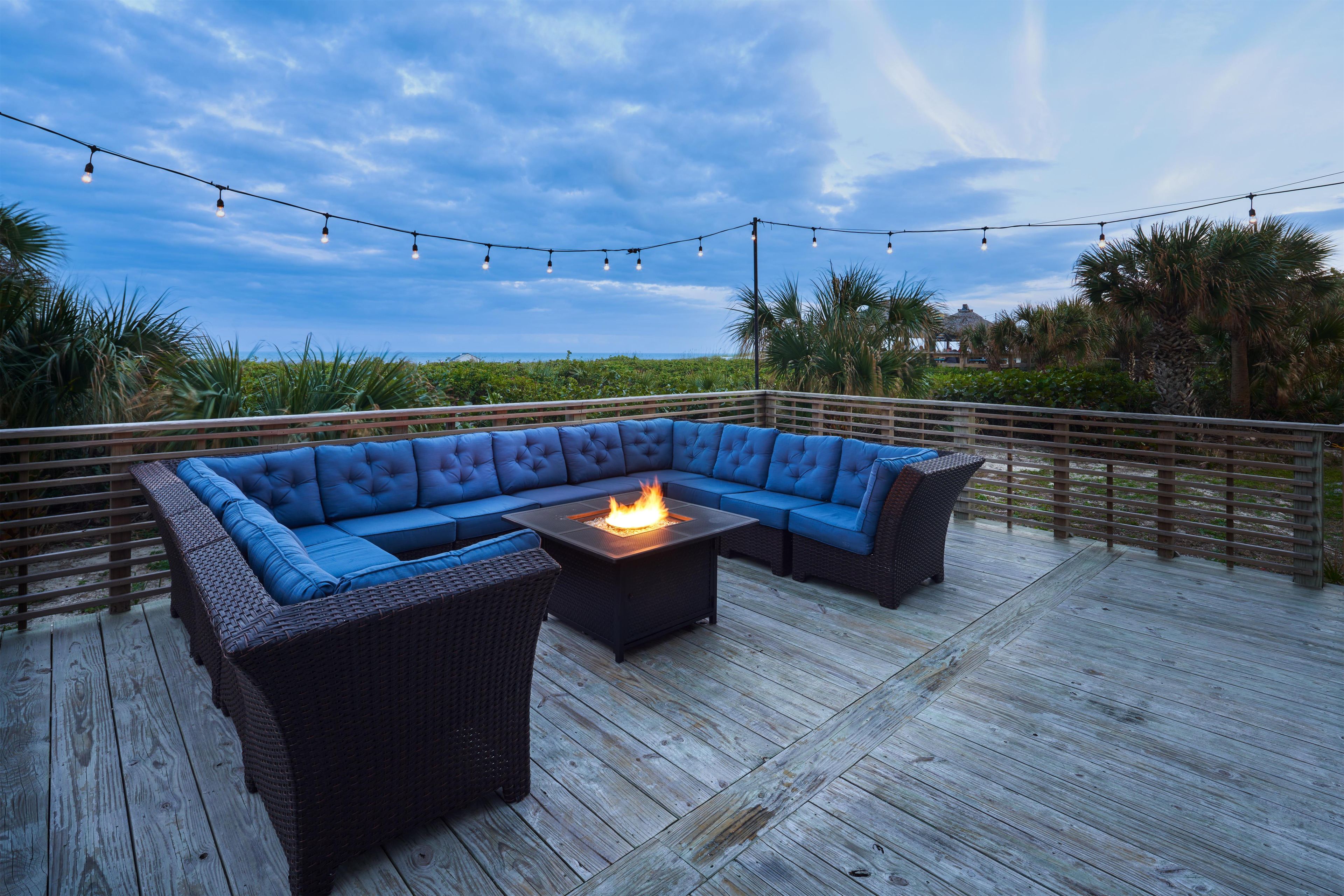 A deck with a blue sectional sofa, a square table with a fire feature, and string lights.