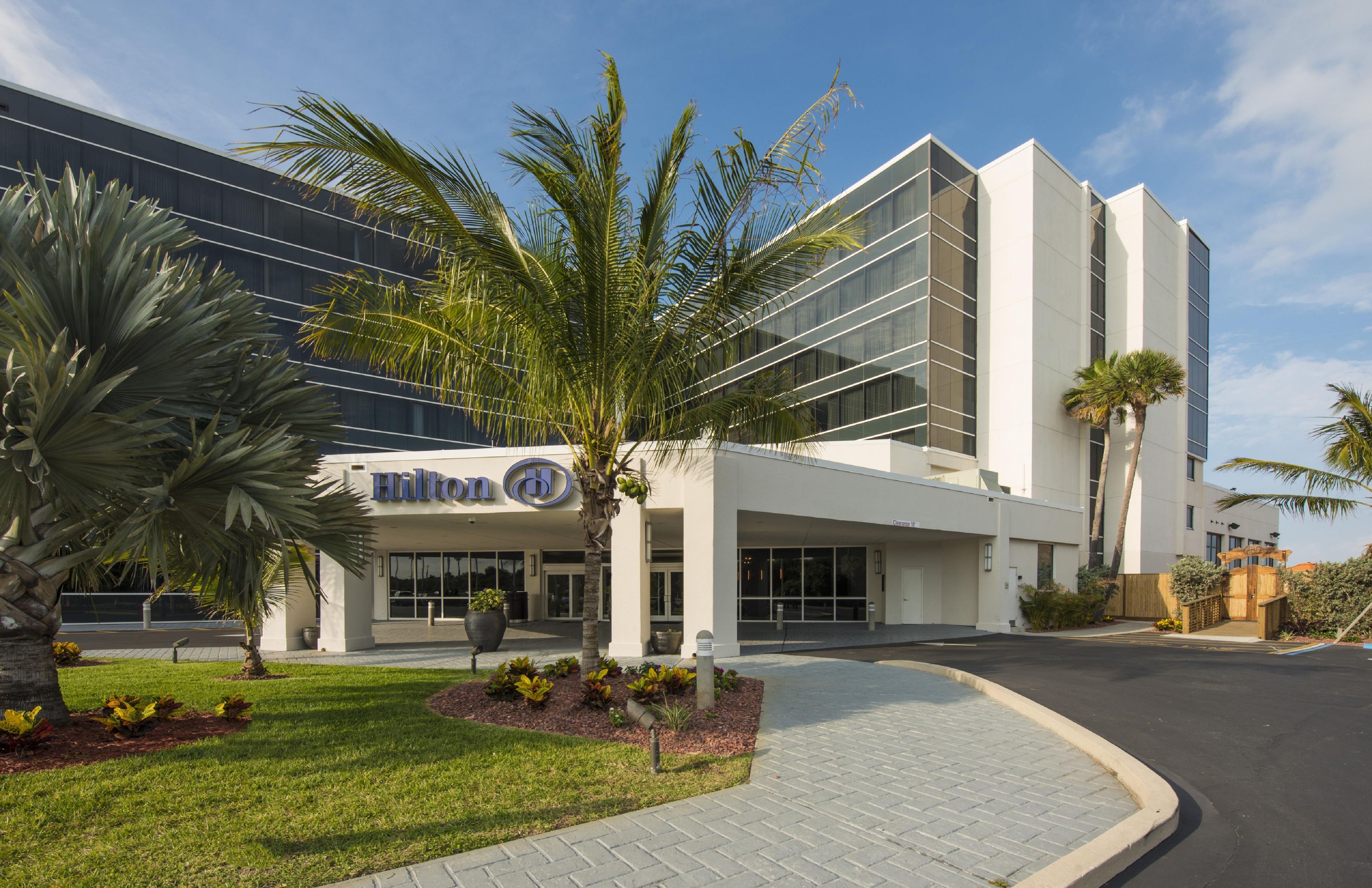 A modern hotel with a glass facade, palm trees, and a well-maintained lawn.