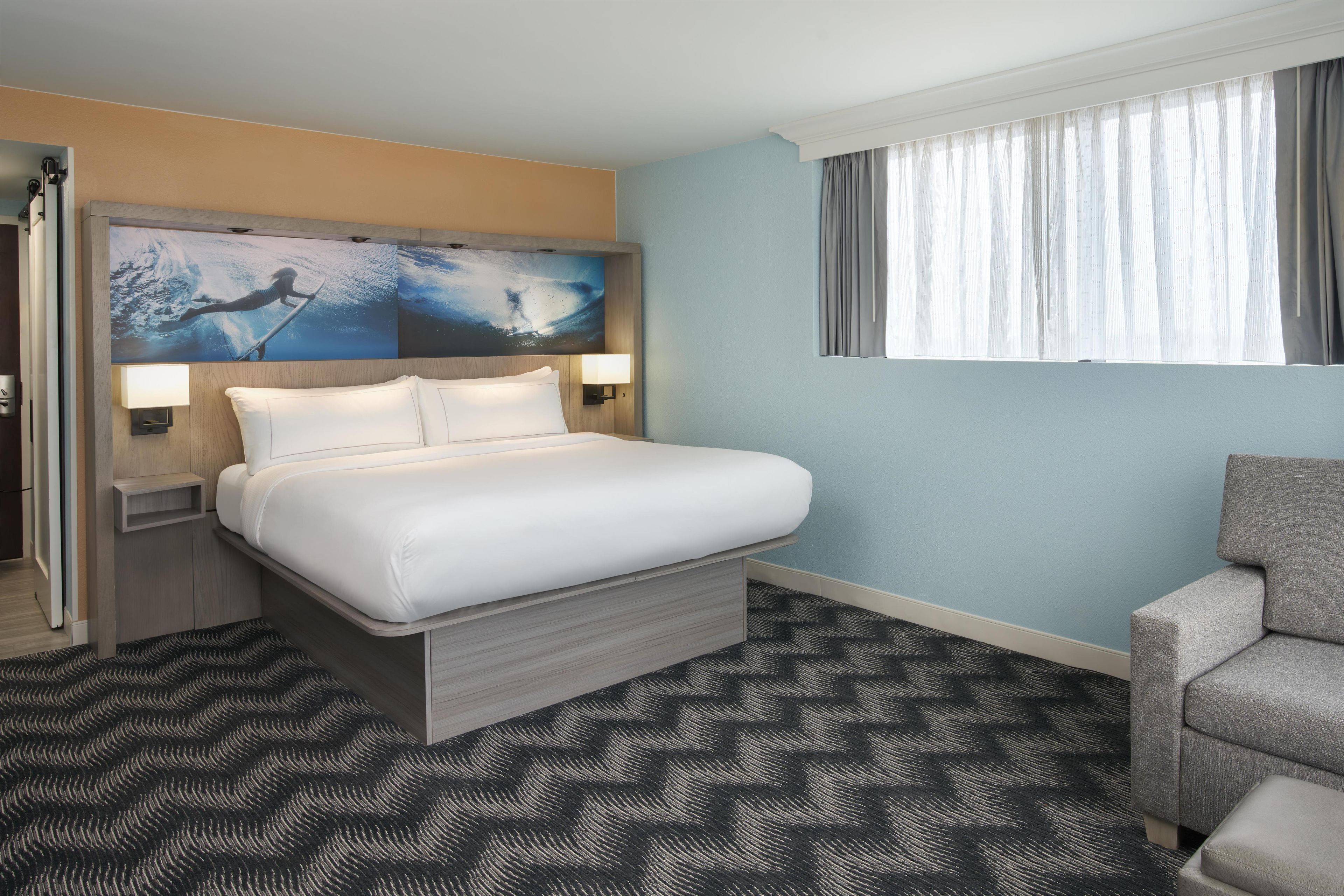 A hotel room with a bed, a chair, a painting, and a window with curtains.