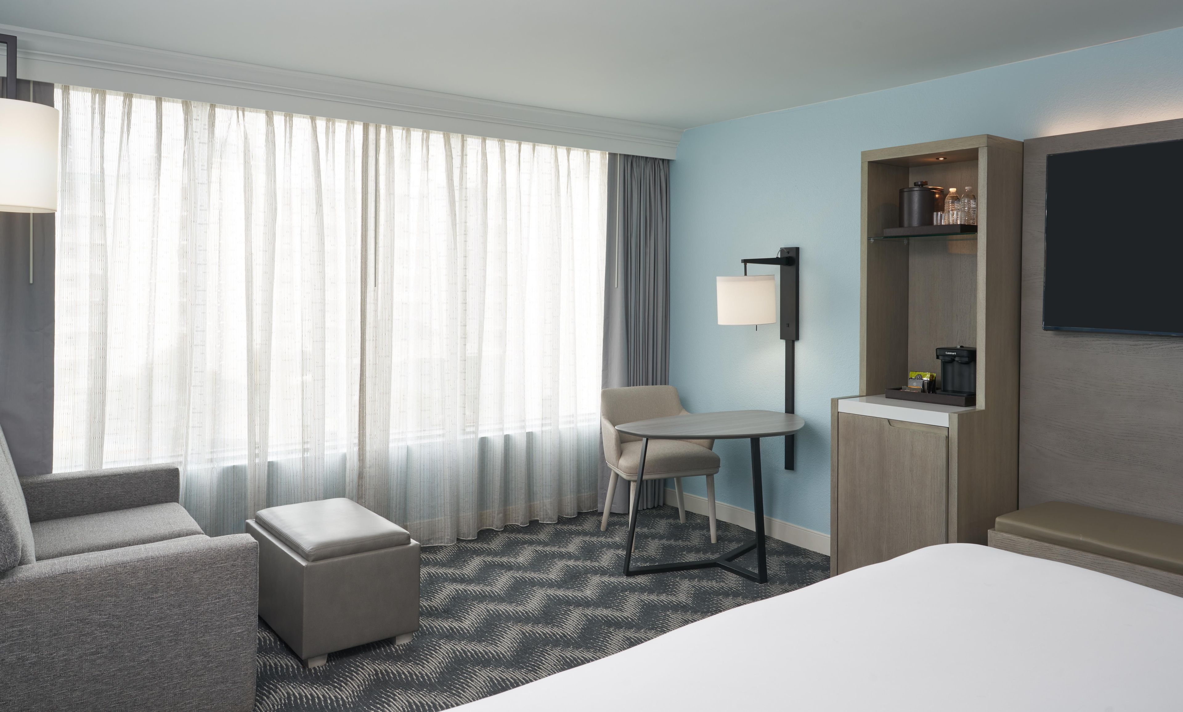 A hotel room with a bed, a desk, a chair, a small table, a sofa, a coffee machine, and a TV.